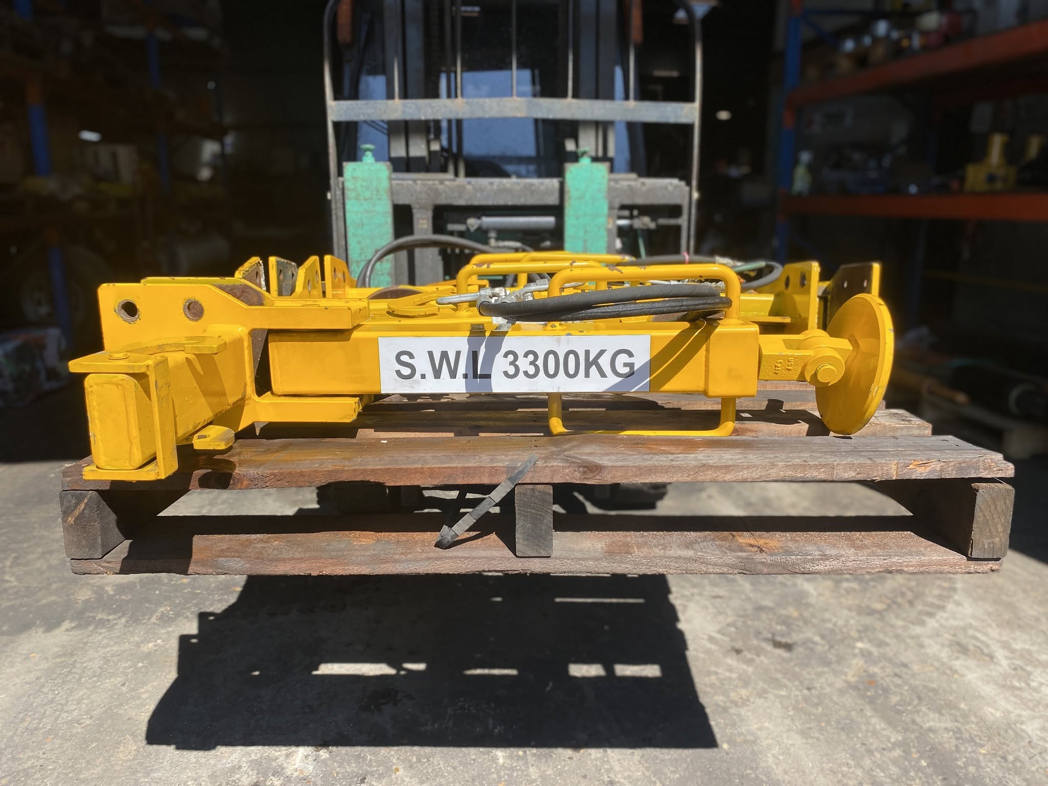Yellow hydraulic lifting attachment with hoses resting on a timber pallet